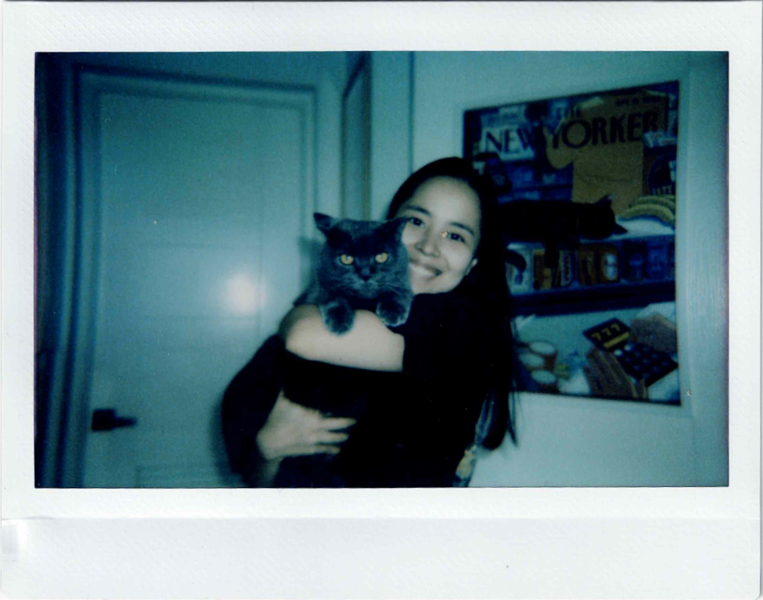 Polaroid of Gabrielle holding her gray cat Bella, with a New Yorker poster in the background